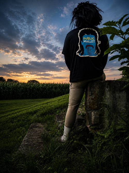 Public Mullet character wearing the First Run tee, standing above a cornfield at dusk with the shirt graphic illuminated against the darkening sky.